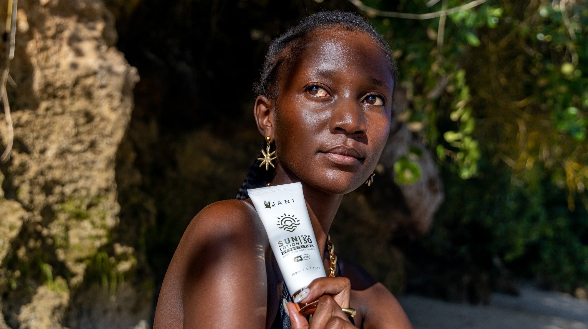 A woman outdoors holding a white tube of ANTI ABRASION SUN PROTECTION 100% OIL, with a rocky, sunlit beach background.