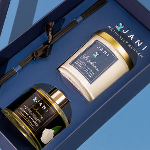 A luxury gift set by JANI, featuring a dark blue box with a glass window lid, containing a cream candle, a dark amber diffuser bottle, and a black leather strap, set against a solid blue background.